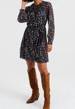 Blumenkleid mit langen transparenten Ärmeln, tailliertem Bund und knielangem Rock in Dunkelblau, verziert mit rosa und goldenen Akzenten. Getragen mit braunen Wildlederstiefeln.