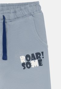 Pantalons à taille élastique bleu clair avec cordon de serrage marine et texte "ROARSOME" imprimé en noir et blanc près de la poche latérale.