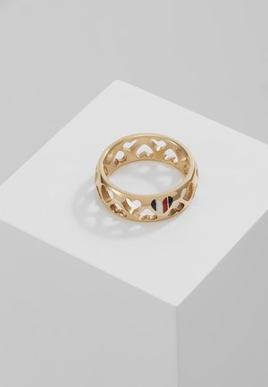 Bague en or avec des cœurs découpés et un cœur rempli de rayures verticales bleues, blanches et rouges, posée sur un piédestal géométrique blanc.