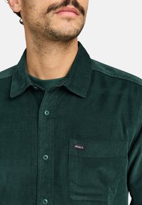 Dark green corduroy shirt with a button-down collar, front pocket, and a small RVCA logo patch. Textured fabric with vertical ribs.