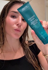 Vrouw met nat haar die een blauwgroene tube van Aveda Botanical Repair versterkende leave-in behandeling dichtbij haar gezicht vasthoudt.