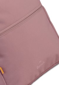 Sac à dos texturé rose avec une poche zippée à l'avant, un accent orange et un logo subtil en bas. Design slim et moderne.