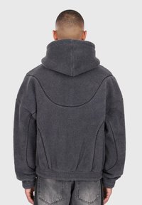 Persona che indossa una giacca in pile grigio scuro con cappuccio, vista da dietro, abbinata a jeans grigi sbiaditi.