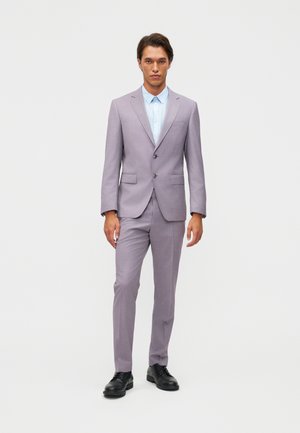 Vyras stovintis dėvintis šviesiai violetinį kostiumą su mėlyna marškinių palaidine ir juodais odiniais batais priešais paprastą šviesią foną.