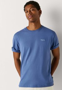 Jeune homme aux cheveux courts et bouclés portant un t-shirt bleu aux manches retroussées, debout devant un fond clair uni.