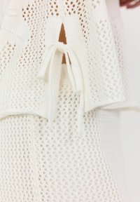 Vitt stickat plagg med en öppen nätdesign, med en knyt-detalj på sidan och en lager-effekt som förbättrar texturen och öppenheten.