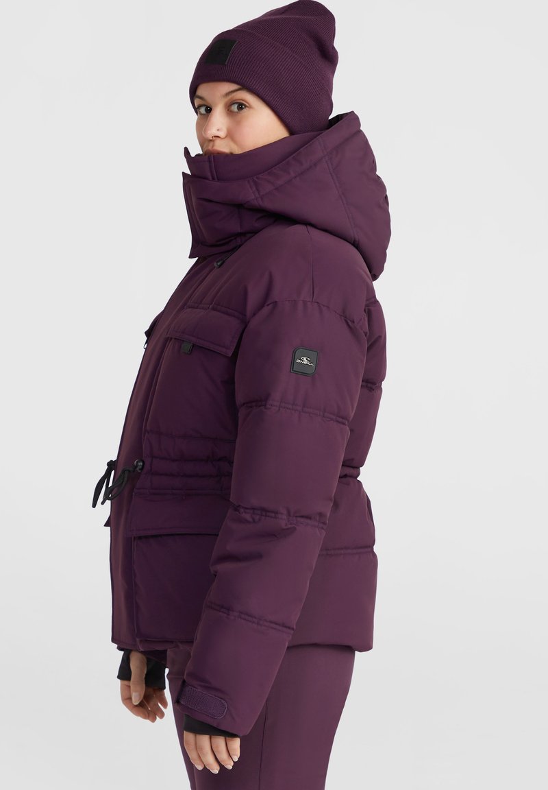 Lila Puffy-Jacke mit hohem Kragen und Kapuze, ausgestattet mit Fronttaschen, verstellbarem Saum und gerippten Bündchen. Getragen mit einer passenden Mütze.