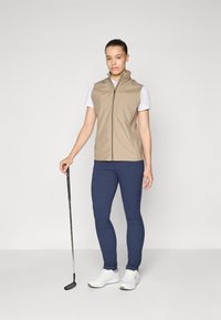 Beige sleeveless vest over a white shirt, paired with navy leggings and white athletic shoes; holding a golf club. Smooth fabric, casual design.
