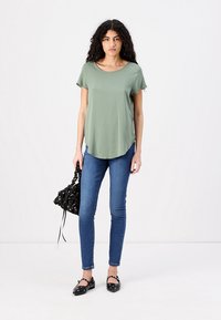 Olive green short-sleeve shirt with a rounded hem, paired with dark blue skinny jeans. Black bag with floral patterns, flat black shoes.
