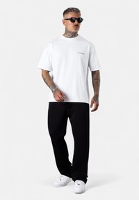 Weißes Baumwoll-T-Shirt mit kurzen Ärmeln, das ein kleines schwarzes Logo auf der Brust trägt, kombiniert mit schwarzen Jogginghosen und weißen Sneakers.