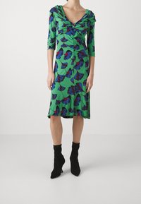 Grön klänning med blå och röda blommönster, rundad halsringning, trekvartslånga ärmar, rynkdetaljer vid midjan och flödande nederkant.