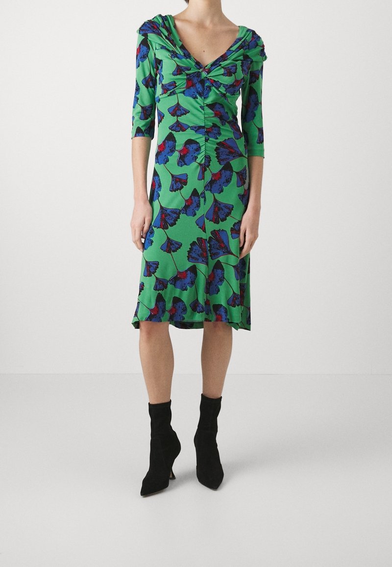 Grön klänning med blå och röda blommönster, rundad halsringning, trekvartslånga ärmar, rynkdetaljer vid midjan och flödande nederkant.