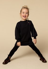 Zwarte oversized sweatshirt met puff mouwen, voorzien van een klein roze hartaccent. Gecombineerd met zwarte leggings en bruine veterschoenen.