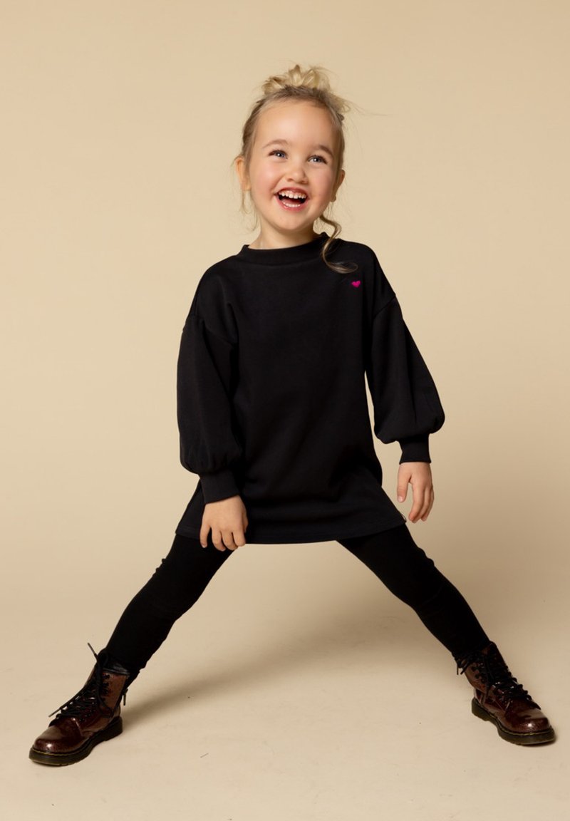 Zwarte oversized sweatshirt met puff mouwen, voorzien van een klein roze hartaccent. Gecombineerd met zwarte leggings en bruine veterschoenen.