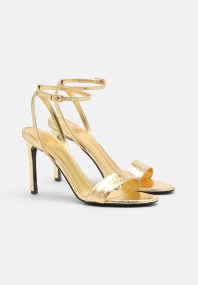 Guld remsandaler med høje hæle, krokodilletryk, åben tå, tynde ankelremme og en sort stilettohæle.