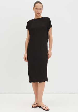 Frau steht, trägt ein schwarzes, knielanges Kleid mit kurzen Ärmeln und schwarze Flip-Flops vor einem einfarbigen weißen Hintergrund.