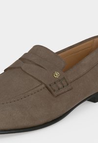 Mocassin en daim marron avec un petit talon noir, détail bride penny et un petit emblème métallique décoratif sur le côté.