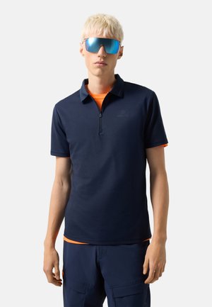 Mand med kort blond hår iført reflekterende blå solbriller, marineblå polo med lynlås, orange undertrøje og marineblå cargobukser mod hvid baggrund.