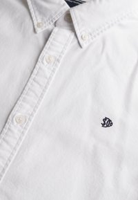 Camisa blanca de botones hecha de algodón; cuenta con un collar puntiagudo, placket frontal con botones y un pequeño logo bordado en azul marino.