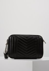 Schwarze Leder-Crossbody-Tasche mit Chevron-Steppdesign, silberner Hardware, Reißverschluss und abnehmbarem Quastenakzent am Riemen.
