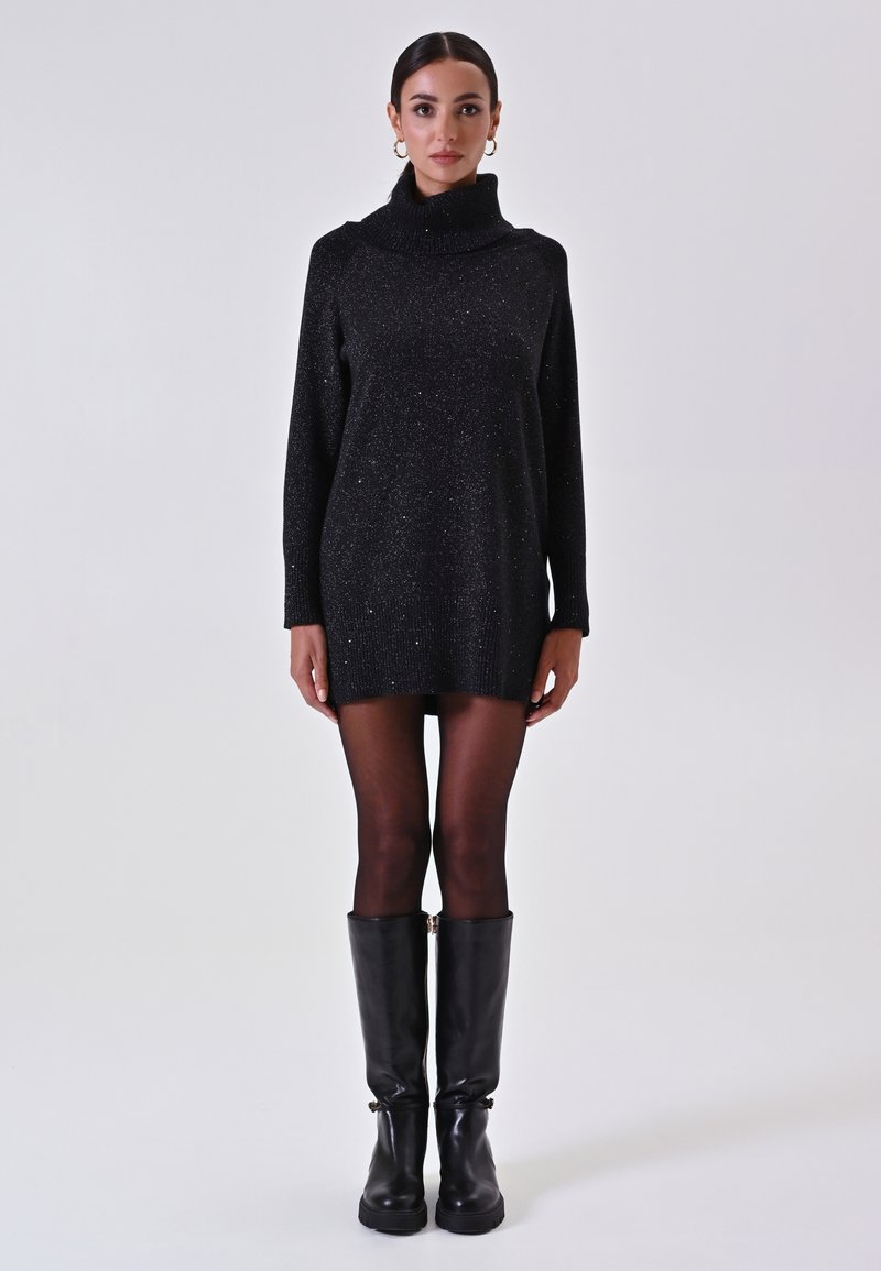 Zwarte sweaterjurk met een hoge kraag, metallic glitters, lange mouwen en een losse fit, gecombineerd met kniehoge zwarte leren laarzen.