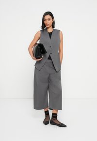 Γκρι αμάνικο γιλέκο με δύο κουμπιά, συνδυασμένο με φαρδιά γκρι παντελόνια. Το μοντέλο κρατά μαύρη γυαλιστερή τσάντα και φοράει μαύρα φλατ παπούτσια με διάφανα καλσόν.