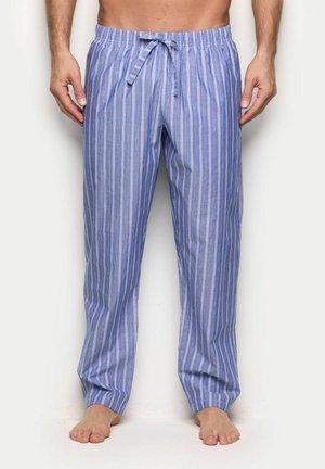 Mann trägt locker sitzende blaue und weiße Pyjamahose mit vertikalen Streifen und Kordelzug, steht barfuß vor weißem Hintergrund.