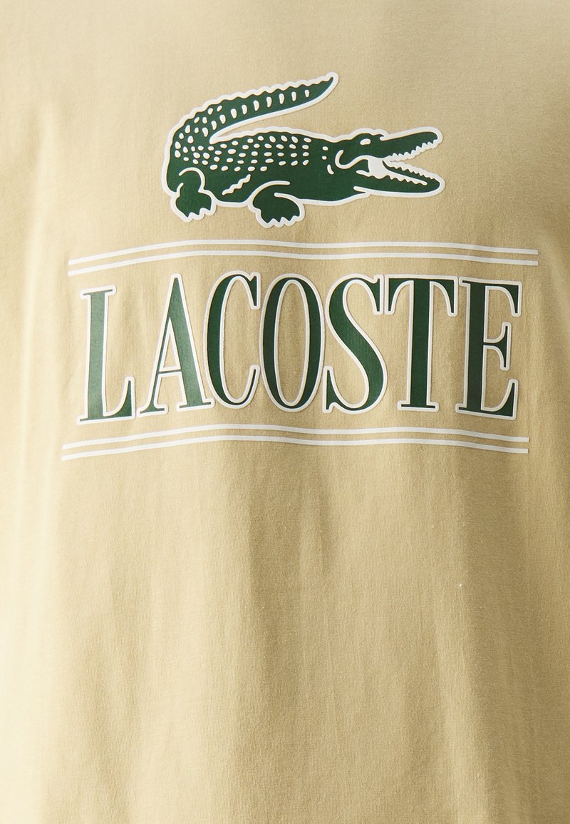 Camiseta de algodón beige con un logo de cocodrilo verde y el texto "LACOSTE" en letras verdes en negrita. Textura suave, mangas cortas y cuello redondo.