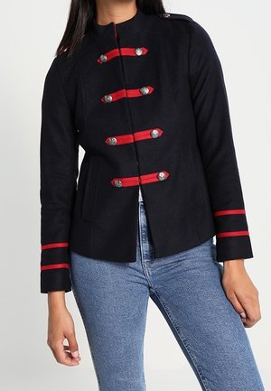 Laivaston villasekoite takki, jossa on punaiset kangaskoristeet ja metallinapit, kaksirivinen malli sekä raidoitusta hihoissa.