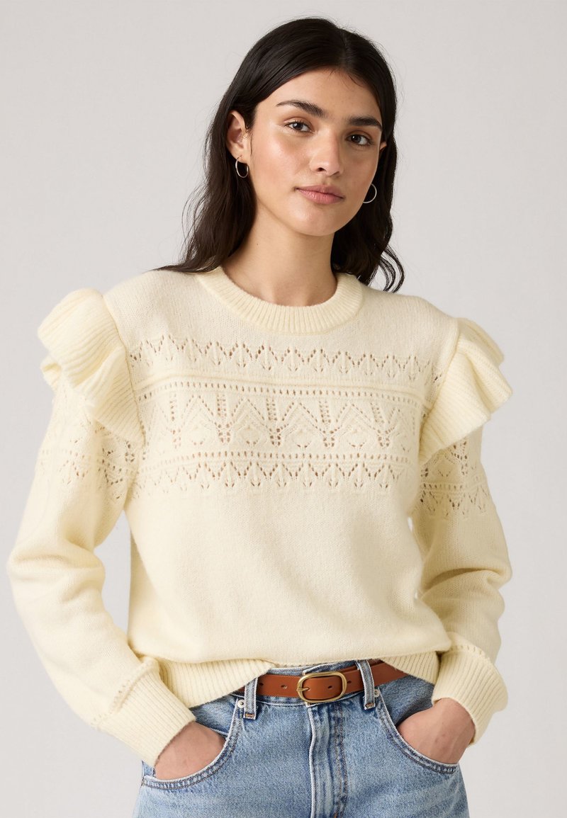 Pull en tricot crème avec des accents de volants aux épaules et un motif ajouré près du col ; associé à un jean en denim taille haute et une ceinture marron.