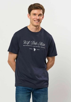 Homme souriant portant un t-shirt bleu marine U.S. Polo Assn. et un jean bleu, debout devant un fond clair uni.
