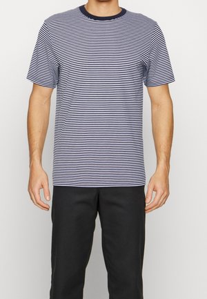 Mann trägt ein kurzärmeliges, marineblau-weiß horizontal gestreiftes T-Shirt und schwarze Hose, steht vor einem schlichten hellen Hintergrund.