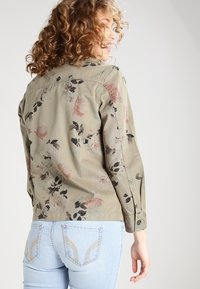 Blommönstrad skjortjacka i olivgrön färg i lätt material. Har knappmanschetter, bakstycke och en böjd fåll, i kombination med blå jeans.