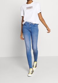 Person som går iklädd blåa skinny jeans, vit T-shirt med grafik, och flerfärgade rutiga sneakers med gula strumpor mot en enfärgad bakgrund.