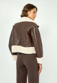 Giacca cropped in pelle marrone con colletto e polsini in pelliccia sintetica bianca; texture liscia; presenta una silhouette aderente e un orlo a contrasto.