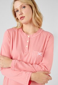 Top rosa a maniche lunghe realizzato in morbido tessuto, con scollatura a bottoni, taschino a pois e cuciture decorative lungo il colletto e le maniche.