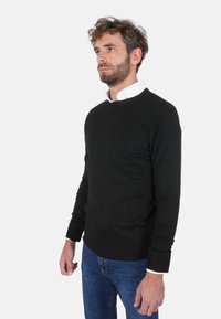 Maglione nero con scollo rotondo, maniche lunghe e polsini a coste, indossato sopra una camicia bianca, abbinato a jeans blu.