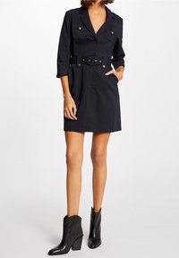 Mörkblå klänning med långa ärmar och en figursydd silhuett. Har två bröstfickor, ett bältat midja och en mjuk tygkänsla. Bärs med svarta ankelboots.