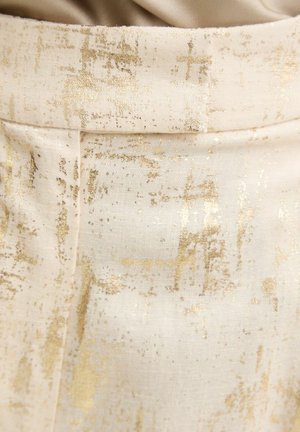 Nahaufnahme eines cremefarbenen Stoffes mit einem goldmetallischen abstrakten Muster und einer Naht, die vertikal nahe der Mitte verläuft.