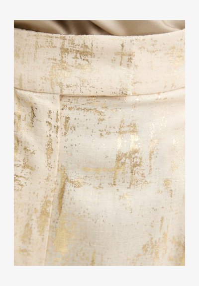 Close-up van crèmekleurige stof met een goud metallic abstract patroon en een naad die verticaal loopt, bijna in het midden.