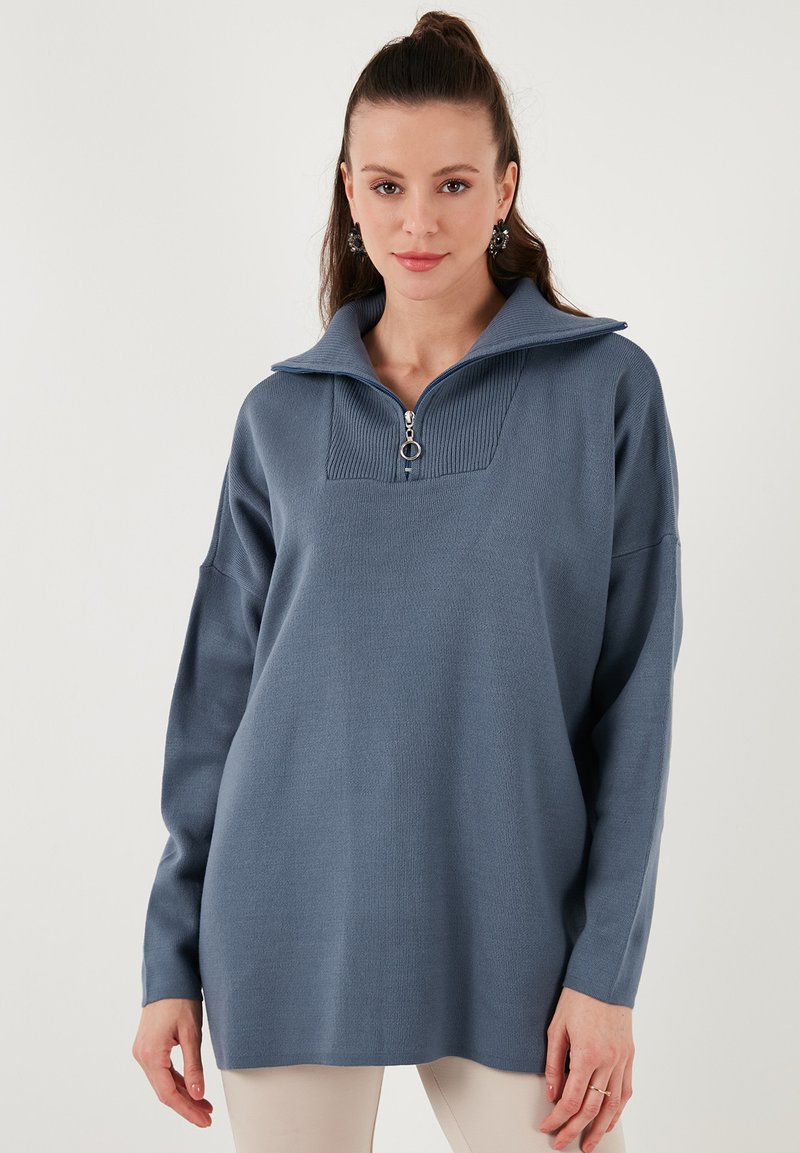 Gaiši zils džemperis ar rāvējslēdzēja kaklu, ribota tekstūra uz apkakles, krītošiem pleciem un brīvu piegriezumu. Gluda auduma, ikdienas stils.