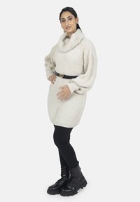 Ljusbeige cowl-neck tröja med ribbad textur, bärs över svarta leggings och kombineras med svarta snörboots. Bälte med hjärtformat spänne.