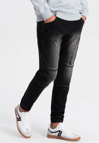 Joggers de mezclilla negra con un ajuste slim, detalles desgastados y puños acanalados. Combinados con zapatillas blancas y grises con suelas marrones.