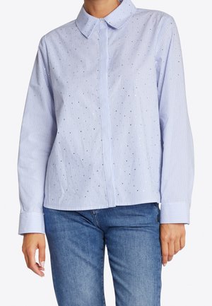 Vrouw in een lichtblauw gestreept overhemd met kleine zilveren studs, gecombineerd met een blauwe spijkerbroek, zichtbaar van schouders tot heupen.