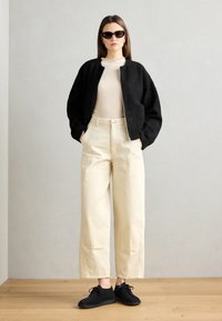 Jachetă texturată neagră, top bej cu dungi, pantaloni largi bej și pantofi negri. Modelul poartă ochelari de soare închiși la culoare, stând în fața unui perete deschis.