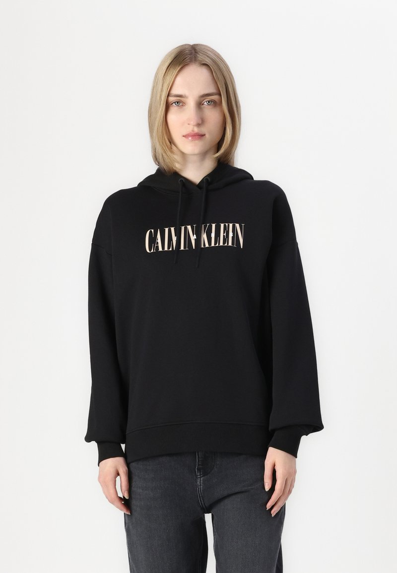 Črna kapuca z ohlapnim krojem in sprednjim logotipom z napisa "CALVIN KLEIN" v bež barvi. Nosite z temnimi, teksturiranimi jeansi.