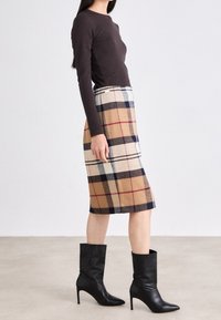 Haut ajusté marron à manches longues associé à une jupe en laine à carreaux de longueur genou dans des tons de beige, marron, marine et blanc. Bottines noires à talons hauts.