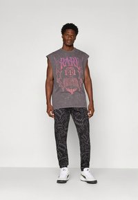 Szürke ujjatlan grafikás póló rózsaszín és fehér nyomattal; mintás fekete nadrág; fehér sportcipő fekete talppal. Egyszínű háttér.