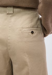 Μπεζ παντελόνι με πίσω τσέπη που κλείνει με κουμπί, συνδυασμένο με καφέ πλεκτό πουλόβερ που είναι μπαλωμένο στη μέση.