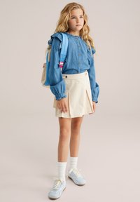 Jeanshemd mit Rüschensäulen, beiger, plissierter Rock, hellblaue Sneakers und weiße Socken. Enthält einen blauen Rucksack mit rosa Akzenten.